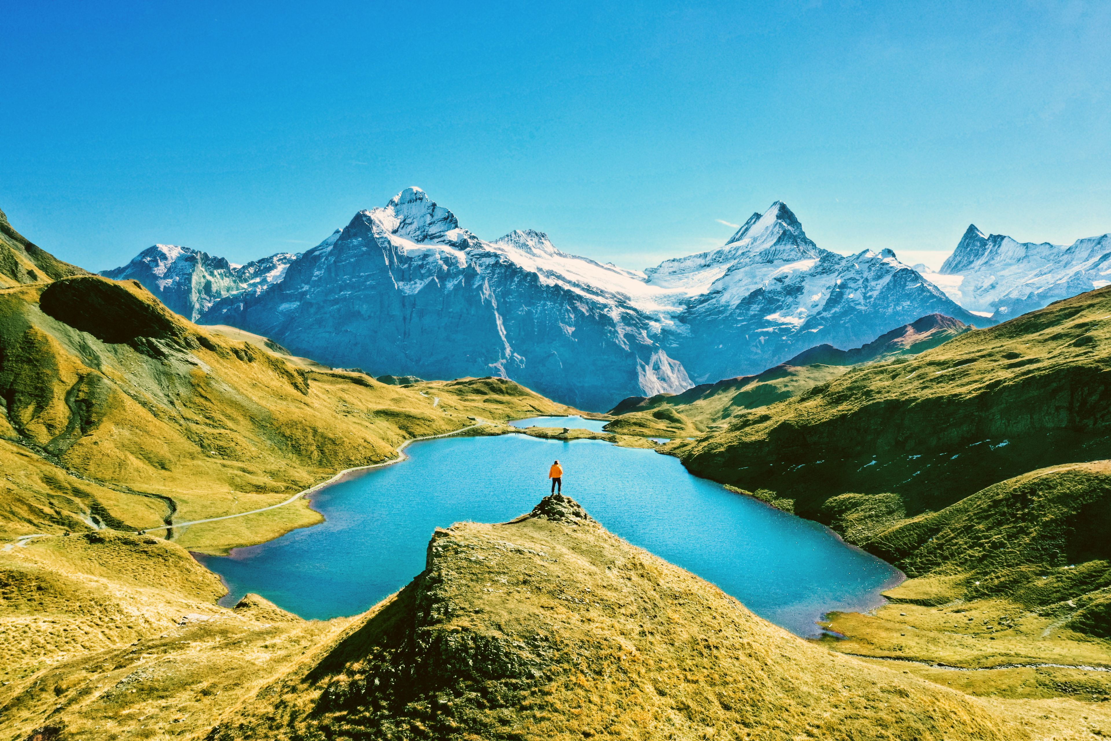 Person in oranger Windjacke spaziert durch eine grüne Bergwiese mit Blick auf einen Bergsee und eine weitläufige Berglandschaft im Hintergrund.